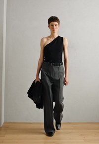 Top negro de un solo hombro combinado con pantalones grises de tiro alto. La modelo sostiene una chaqueta negra; diseño minimalista sobre un suelo de madera clara.