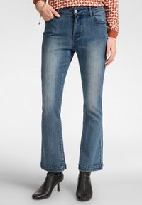 Jeans de mezclilla azul de corte acampanado con una cintura de altura media, cinco bolsillos y un acabado desgastado. Llevados con botas de tacón negras hasta el tobillo.