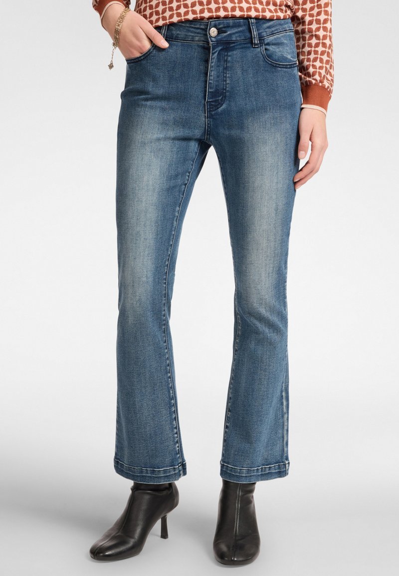 Jeans de mezclilla azul de corte acampanado con una cintura de altura media, cinco bolsillos y un acabado desgastado. Llevados con botas de tacón negras hasta el tobillo.