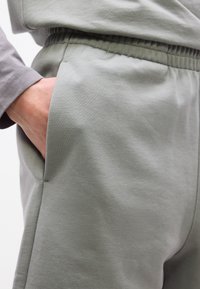 Άτομο που φοράει αθλητική φόρμα με ελαστική μέση σε ανοιχτό γκρι χρώμα, με το χέρι στην πλαϊνή τσέπη, εμφανής τμήμα από το αντίστοιχο μακρυμάνικο πουκάμισο.