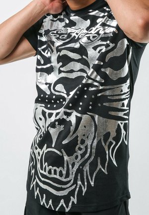 Person iført en sort T-shirt med et stort sølvfarvet abstrakt dyreansigt-design og teksten "Ed Hardy" nær halsudskæringen.