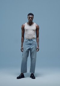 Beige tanktop, lichtblauwe oversized jeans en zwarte schoenen. Neutrale achtergrond, gericht op productdetails en pasvorm.