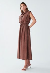 Brown, sleeveless maxi dress with a button-up front, drawstring waist, and white polka dots. Paired with white heeled sandals.