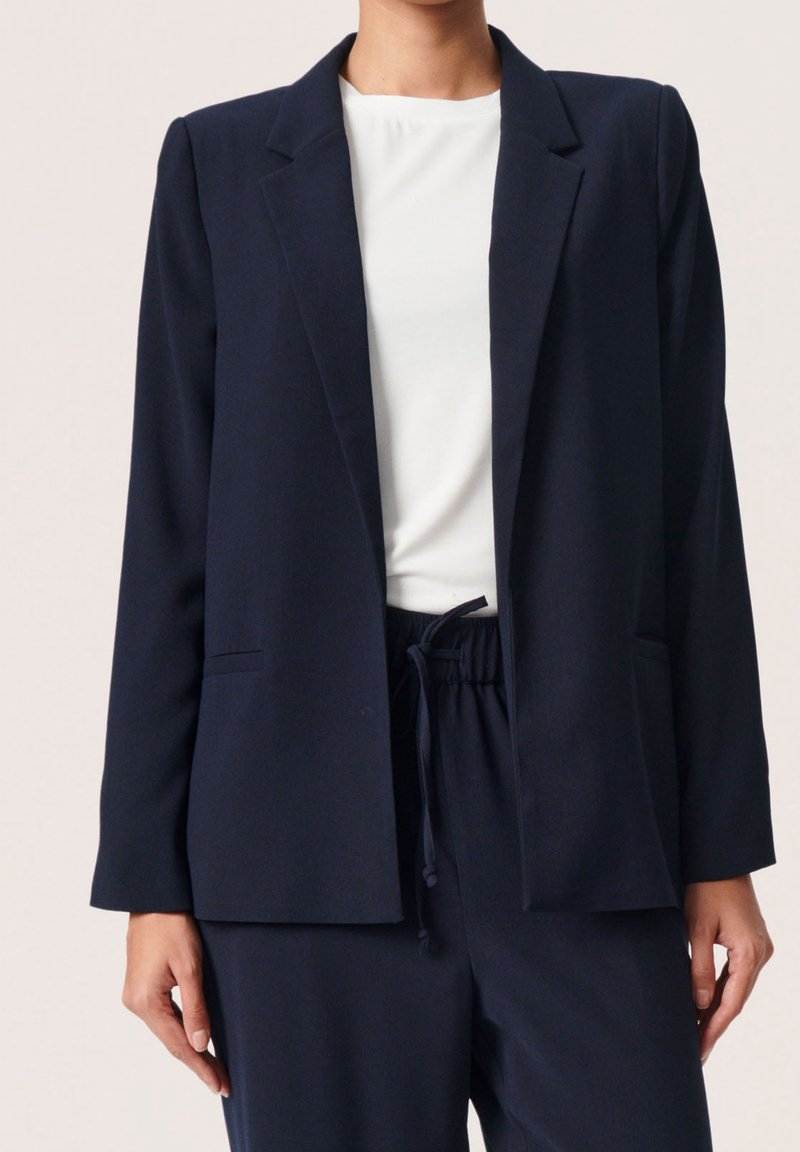 Námořnický blazer s strukturovaným designem, dlouhými rukávy, klopami a dvěma předními kapsami. Nosený přes bílou košili a tmavé kalhoty.