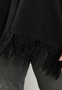 Svart fransad stickad plagg som bärs över mörkgrå jeans, som visar nedre delen av kroppen och högerhanden med ljusrosa nagellack.