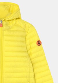 Gul pufferjakke med hætte, der har en quiltet tekstur og et rundt orange logo badge på venstre ærme. Sølv lynlås med et orange greb.