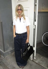 Žena s blond vlasmi, ktorá nosí okuliare, bielu polokošeľu, široké tmavé džínsy, drží čiernu kabelku a stojí vo dverách na ulici.