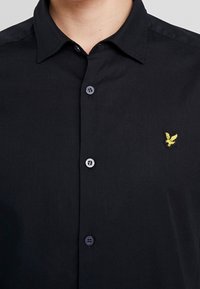 Zwart overhemd met knopen, gemaakt van gladde stof. Drie knopen zichtbaar, met een klein geel geborduurd logo op de linkerborst.