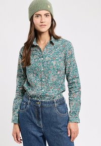 Türkisfarbendes, floral bedrucktes Hemd mit Knopfleiste und langen Ärmeln, kombiniert mit hoch taillierten blauen Jeans und einer hellgrünen Strickmütze.
