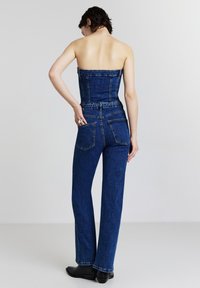 Tuta in denim blu con design senza spalline, corpino aderente, pantaloni a gamba larga e due tasche posteriori. Materiale testurizzato e casual.