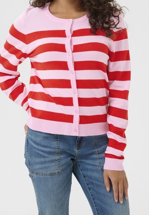 Woman wearing pink and red horizontal striped buttoned cardigan with blue jeans, standing with one hand on hip and the other by side.