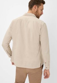 Beige long-sleeve shirt with a collar, button cuffs, and a slightly elongated back hem. Made from textured, lightweight fabric.