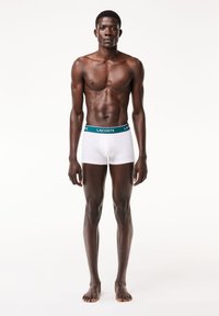 Boxeri albi cu un waistband verde cu logo Lacoste, design ajustat și textură netedă, modelați de o persoană pe un fundal alb.