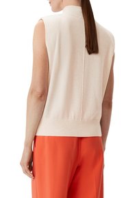 Miękki beżowy sweter bez rękawów z wysokim kołnierzem, prążkowanym pasem i centralnym detalem szwu, w połączeniu z jasnopomarańczowymi spodniami.