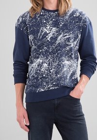 Sudadera azul marino con un patrón de salpicaduras blancas, mangas largas, puños y dobladillo acanalados, hecha de una tela texturizada. Pantalones vaqueros oscuros usados debajo.