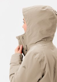 Beige jasje met een capuchon, voorzien van een ritssluiting aan de voorkant, een verstelbare hals en logokleuren. Glad textuur, lichtgewicht materiaal zichtbaar.