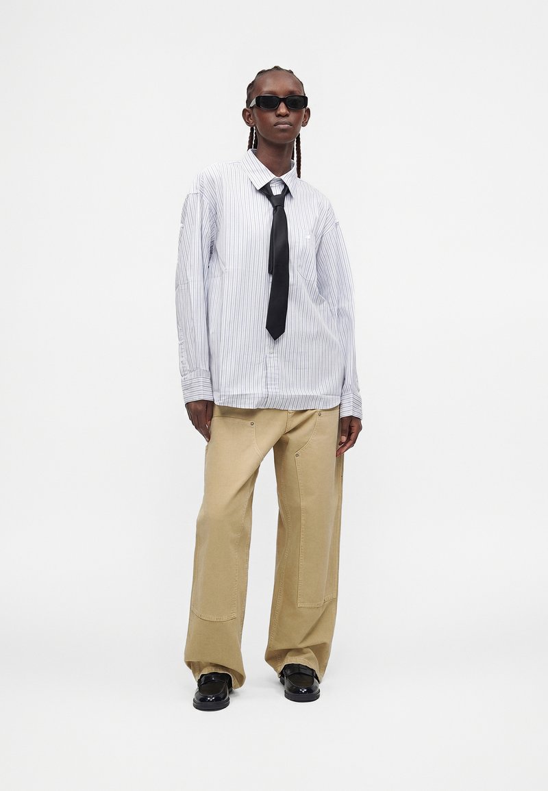 Persona che indossa occhiali da sole neri, camicia bianca a righe, cravatta nera, pantaloni beige a gamba larga e scarpe nere, in piedi su uno sfondo bianco uniforme.