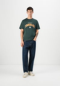 Camiseta de algodón verde con el logo de "Timberland" en naranja, combinada con unos vaqueros de denim azul sueltos y zapatillas crema. Conjunto sencillo y casual.