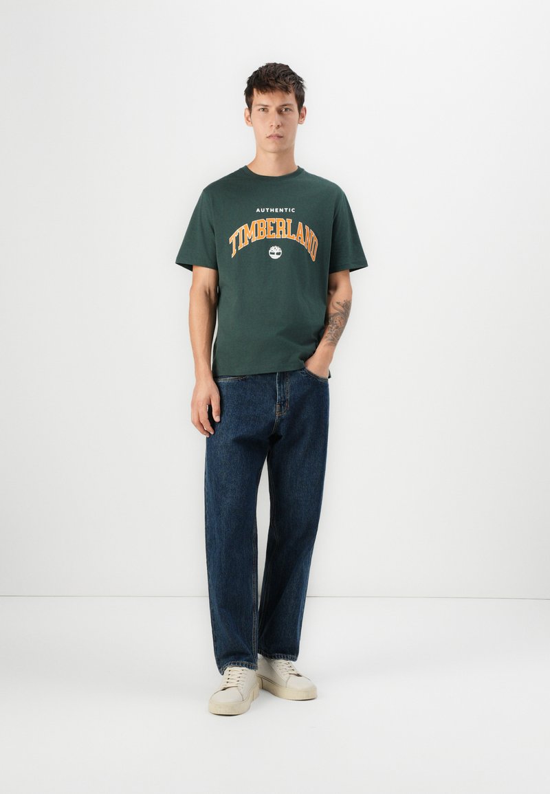 T-shirt in cotone verde con logo "Timberland" arancione, abbinato a jeans in denim blu larghi e sneakers beige. Outfit semplice e casual.