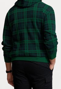 Zelená kostkovaná mikina s kapucí a tmavými doplňky, s kapucí, žebrovanými manžetami a lemem, vyrobená z měkkého materiálu. Pod ní jsou vidět jednoduché černé kalhoty.