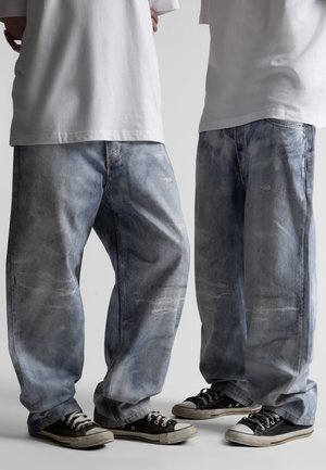 Două persoane care poartă blugi albaștri largi și decolorați, tricouri albe largi și adidași negri cu talpă joasă, stând una lângă alta.