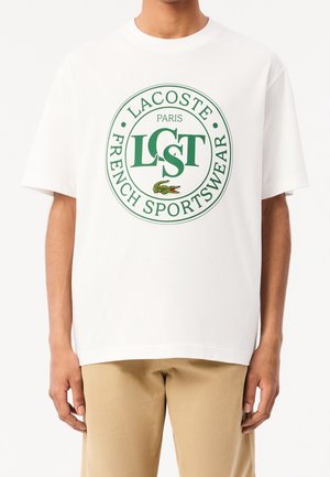 Weißes Baumwoll-T-Shirt mit rundem grünem Logo, auf dem der Text "LACOSTE PARIS FRENCH SPORTSWEAR" und eine Krokodil-Grafik in der Mitte abgebildet sind.