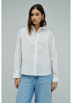 Jeune femme aux longs cheveux noirs portant une chemise blanche à boutons et un jean bleu clair, devant un fond uni gris clair.