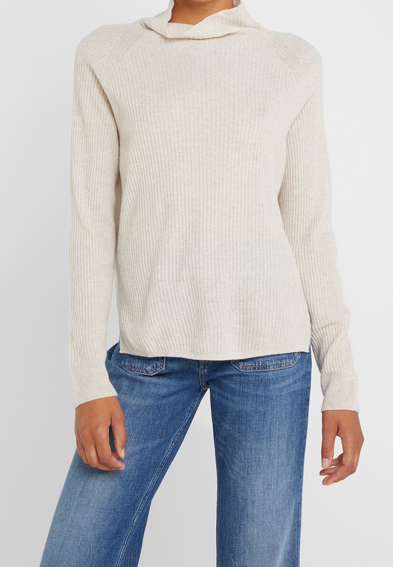 Persoon in een beige geribbelde trui met lange mouwen en blauwe denim jeans, staande tegen een plain witte achtergrond.
