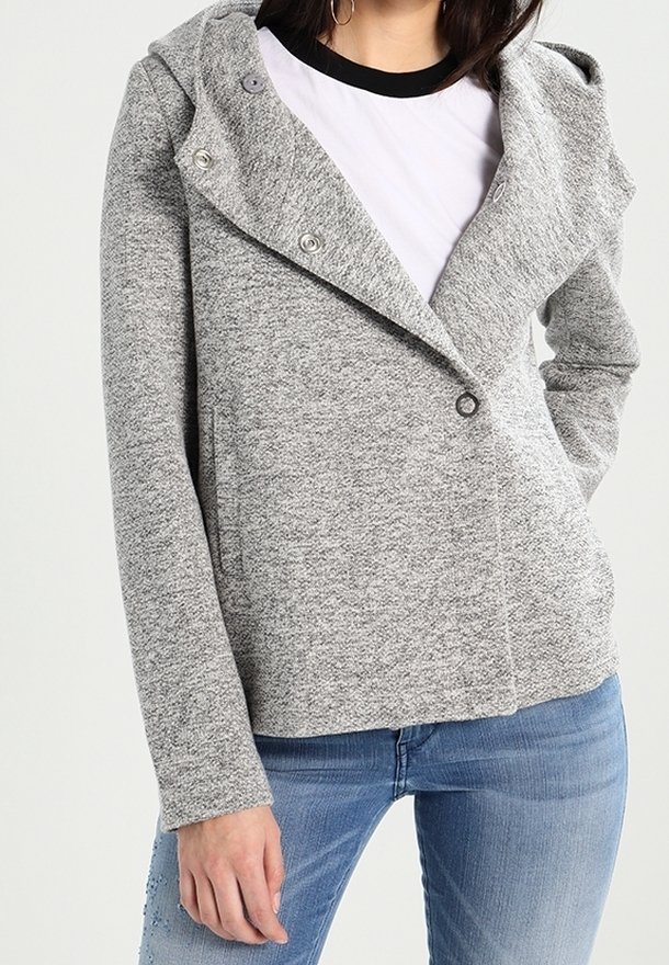 Femme portant une veste à capuche gris chiné par-dessus un t-shirt blanc et un jean bleu, avec une main dans une poche.