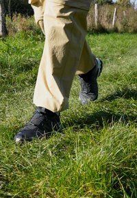 Black outdoor shoes with textured surfaces and laces, worn with light-colored pants, walking on green grass in a natural setting.