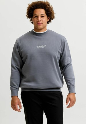 Jeune homme aux cheveux bouclés portant un sweat-shirt gris JJ Studios et un pantalon noir, debout devant un fond uni.