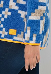 Niebieska bluza polarowa z geometrycznymi wzorami w kolorze beżowym i białym. Ma pomarańczowe wykończenia oraz małą żółtą metkę z nazwą marki przy krawędzi.