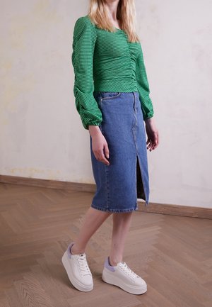 Kvinde, der står indendørs iført en grøn rynket top, knælængde denimnederdel med frontsplit og hvide platformsneakers.