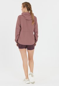 Lichtgewicht mauve regenjack met een capuchon, lange mouwen, elastische manchetten en een gebogen zoom, gedragen over donkere shorts en witte sneakers.