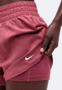 Άτομο με αθλητικά σορτς Nike σε ροζ χρώμα κρατά το ύφασμα στο στρίφωμα, δείχνοντας το λευκό λογότυπο της Nike κοντά στο κάτω άκρο.