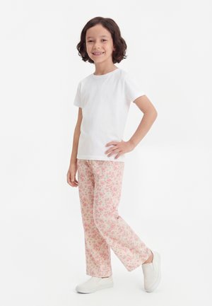 Dieťa s krátkymi tmavými vlasmi stojí a usmieva sa, oblečené v jednobielom tričku, ružových kvetovaných nohaviciach a bielych slip-on topánkach.