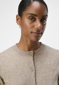 Lysebrun strikket cardigan med rund halsutringning, to knapper og ribbestrikket tekstur. Modellen vises i en nærbilde med kort hår og hoopsmykke.