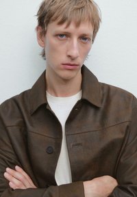 Giacca di pelle marrone con chiusura a bottoni, caratterizzata da un colletto a punta e una texture sottile, indossata sopra una camicia di colore chiaro.