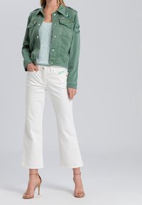 Veste en denim vert avec plusieurs poches, jeans blancs à jambes larges et sandales à talons beige à lanières. Le haut est un débardeur texturé bleu clair.