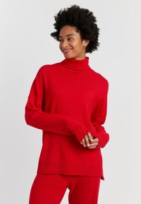 Pullover a collo alto rosso, texture a maglia, vestibilità rilassata, maniche lunghe, spacchi laterali, abbinato a pantaloni rossi coordinati.