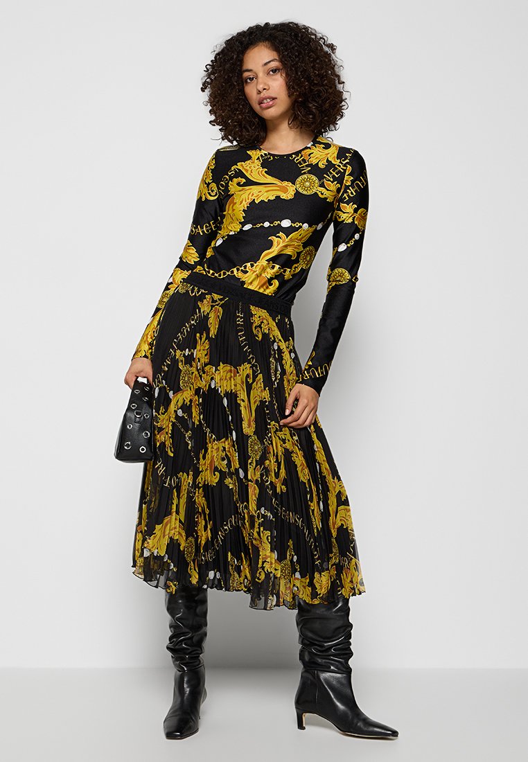 Femme posant dans un haut à manches longues noir et jaune à motifs et une jupe midi plissée, tenant un sac à main noir et portant des bottes noires.