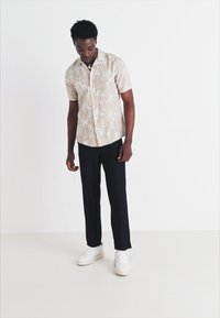 Short-sleeve beige button-up shirt with a palm tree pattern, paired with black trousers and white sneakers. Model stands facing down.