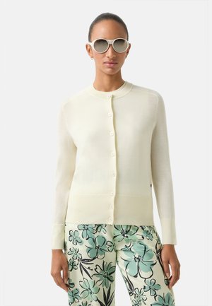 Lys gul cardigan med rund hals og knaplukning, kombineret med myntegrønne bukser med blomsterprint, der har en glat tekstur.