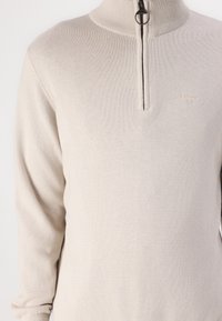 Beige gestrickter Pullover mit einem halben Reißverschluss am Kragen, der eine dezente Textur und ein kleines Markenlogo auf der Brust aufweist. Mit langen Ärmeln.