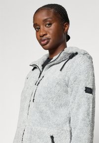 Hoodie di colore grigio chiaro con texture in pelliccia, dotato di zip frontale, cappuccio regolabile e punti di estrazione neri, con tasche laterali e un'etichetta con logo.