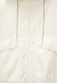 Leichte, cremefarbene Jacke mit Frontreißverschluss, Kordelzügen und zwei großen Taschen. Besitzt einen strukturierten Stoff mit glatter Oberfläche.