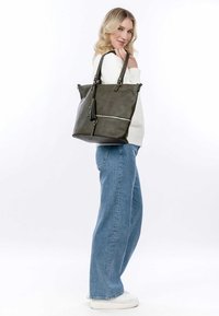 Donna che indossa un maglione bianco e jeans blu, con una grande borsa a tracolla grigio scuro, in piedi contro uno sfondo bianco.