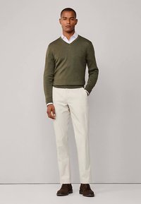 Olivgrön stickad tröja med V-ringning, kombinerad med krämfärgade byxor. Bruna skor kompletterar outfiten. Rena linjer och figursydd design.