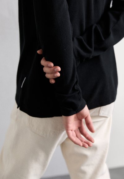 Schwarzes Langarmshirt mit taillierten Ärmeln, getragen zu hellen Hosen. Eine Hand ist hinter dem Rücken platziert, was eine lässige Haltung zeigt.