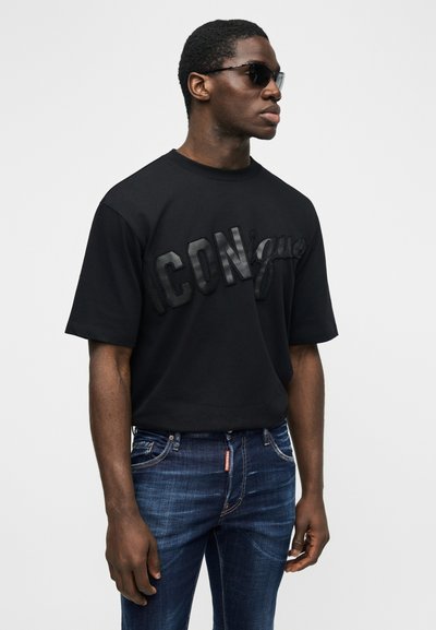 Homme portant des lunettes de soleil noires, un t-shirt noir oversize avec le texte "ICON" et un jean bleu foncé, debout devant un fond clair uni.